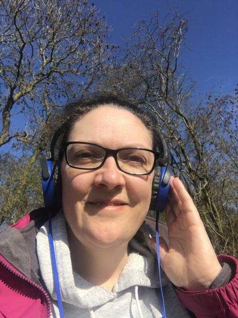 Picture of a woman with brown hair, wearing blue headphones, black glasses and a pink coat